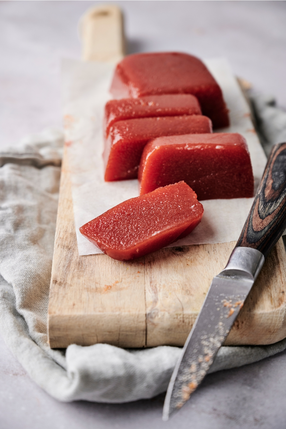 A sliced block of guava paste is on a wooden serving board lined with parchment paper. A wooden handled knife is resting on the board, as well.