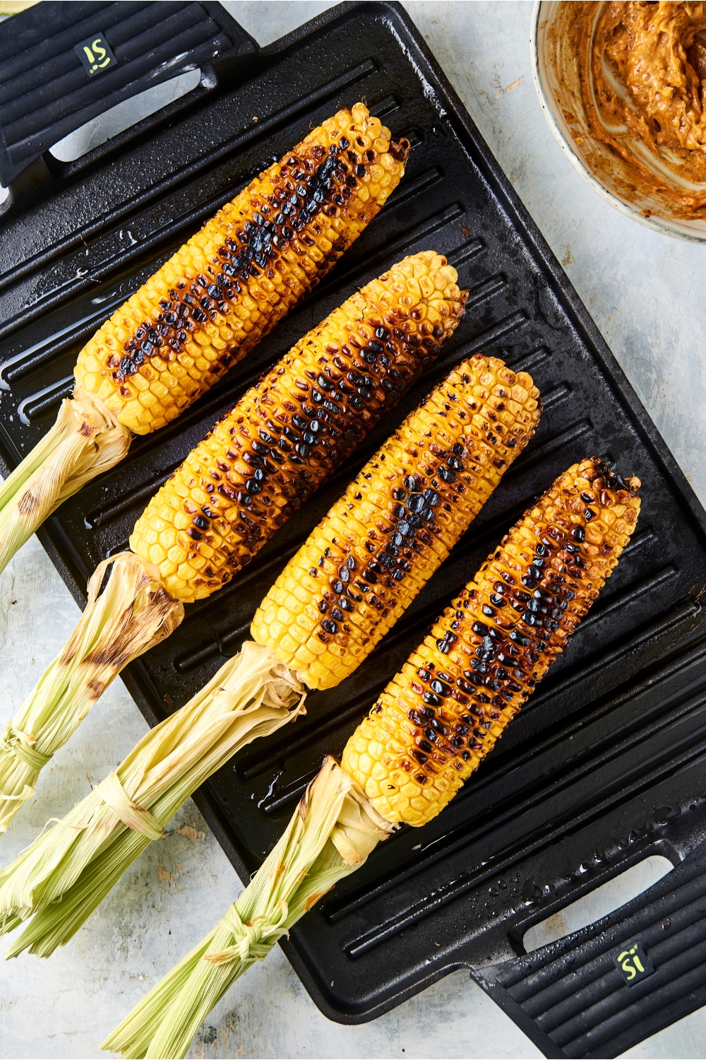 Four ears of corn are on a grill pan. The corn has been charred on each side.