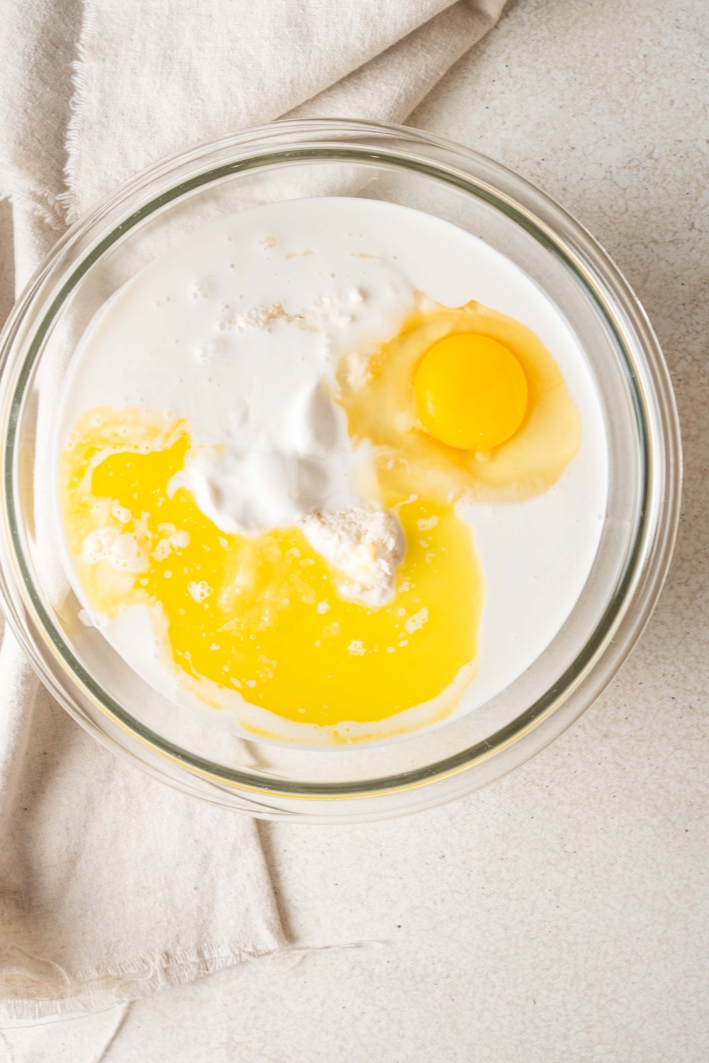 A glass bowl holds cassava cake ingredients. There is a large egg yolk and melted butter sitting atop the ingredients.