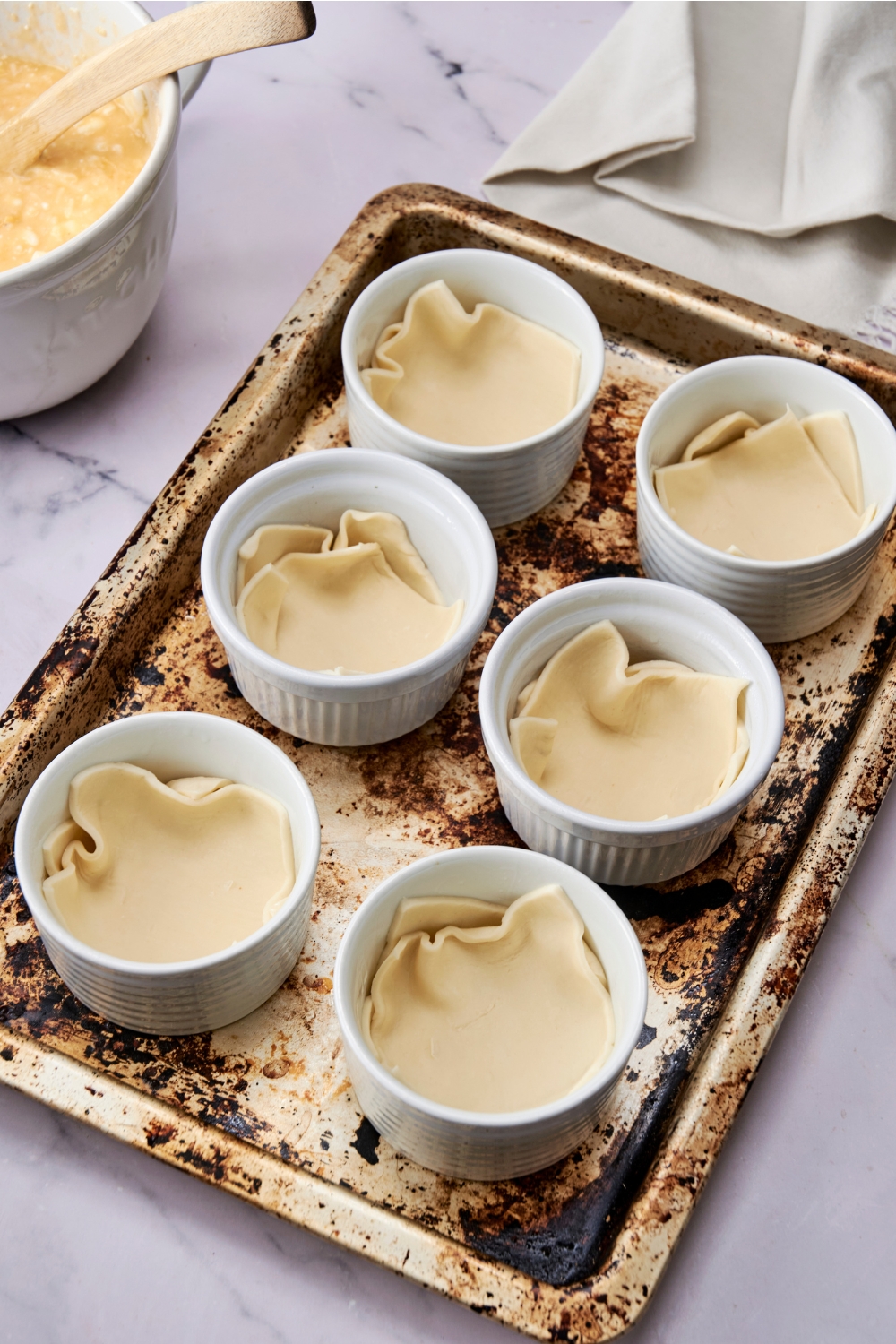 Six ramekins are lined up in two rows on a baking tray. There are two pieces of puff pastry in each ramekin.