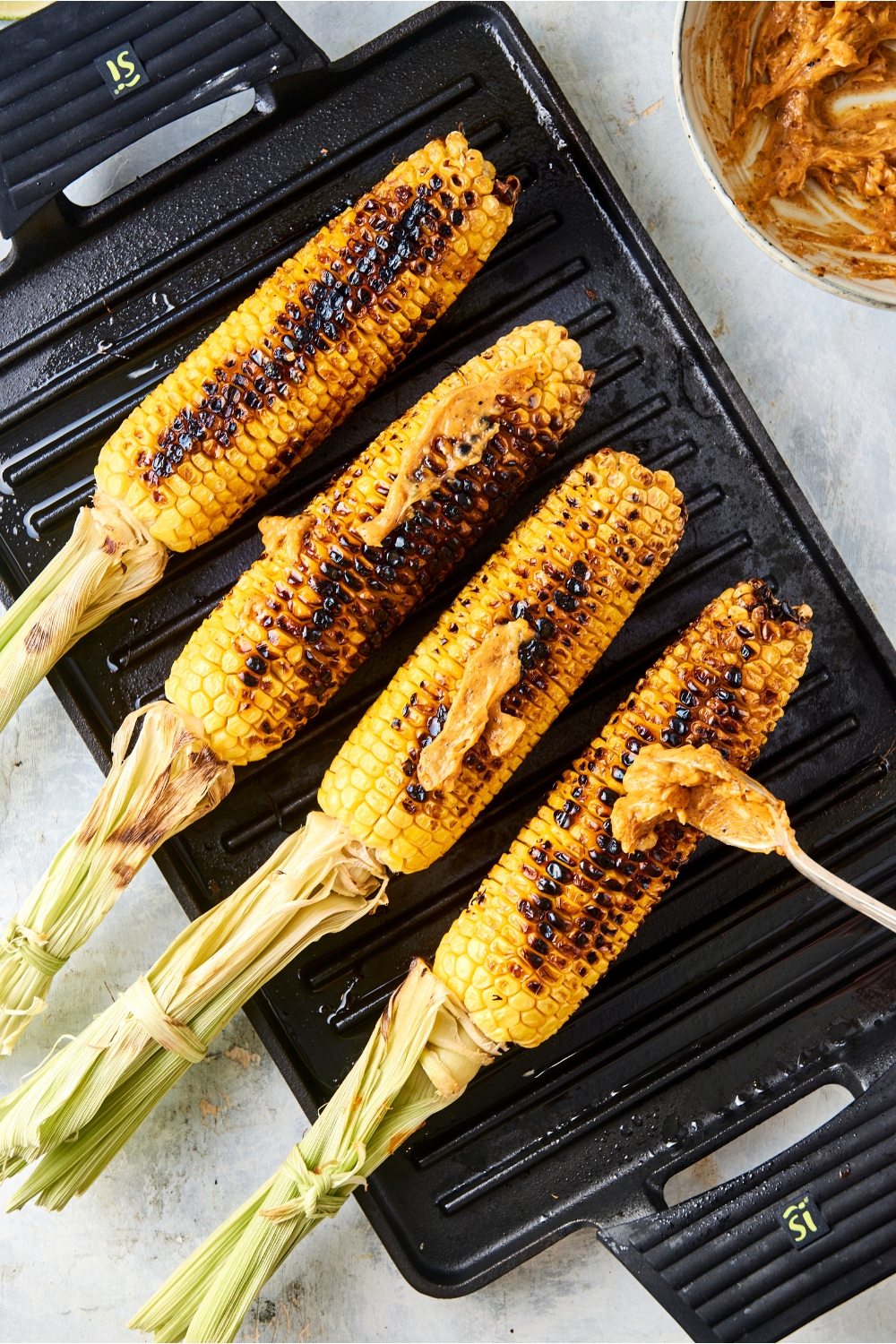 Someone spreads cajun seasoned butter over four cobs on corn on a grill pan.