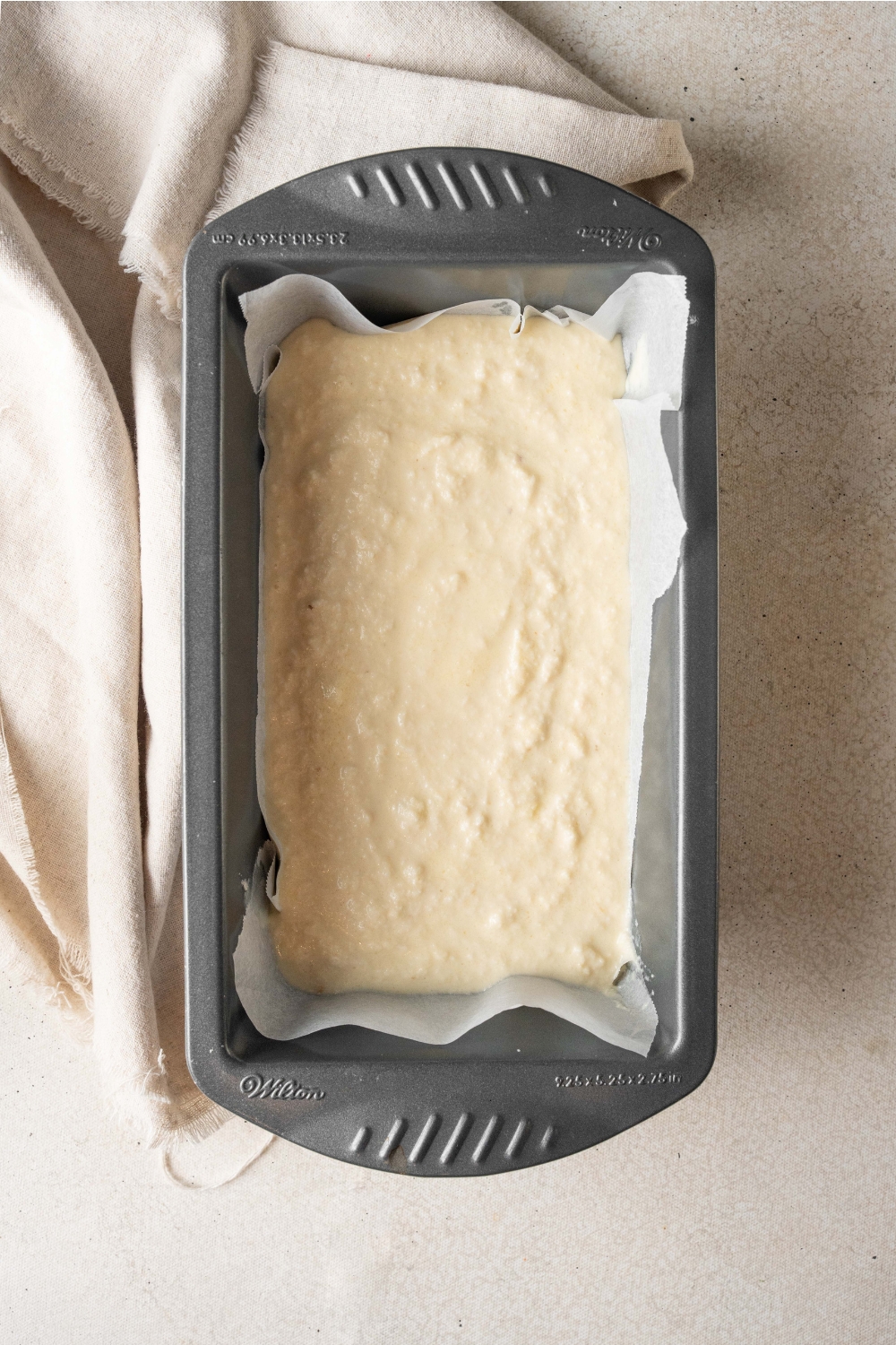 A black loaf pan is lined with parchment paper and is full of cassava cake batter.