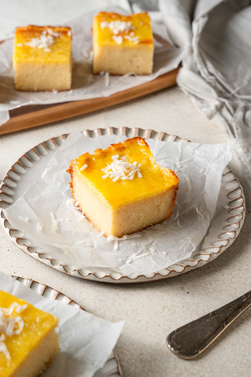 A square piece of cassava cake sits on a parchment paper lined plate. The cake is garnished with shredded coconut.