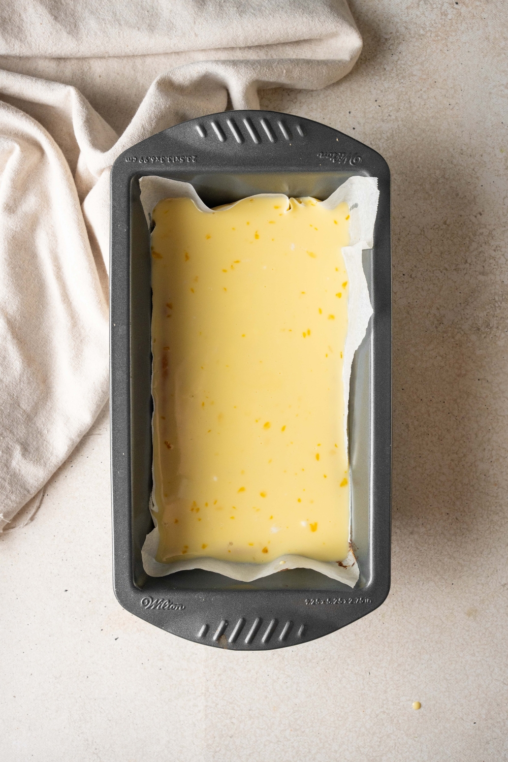 A black loaf pan is lined with parchment paper and holds a baked cassava cake. The cake is topped with uncooked coconut topping.