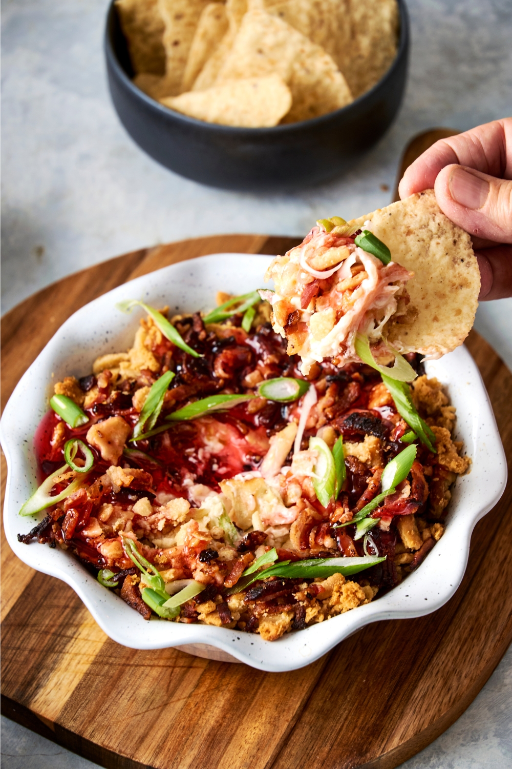 A serving bowl with captain rodney's dip garnished with captain rodney's pepper sauce and chopped green onion. A tortilla chip is being dipped in the bowl.