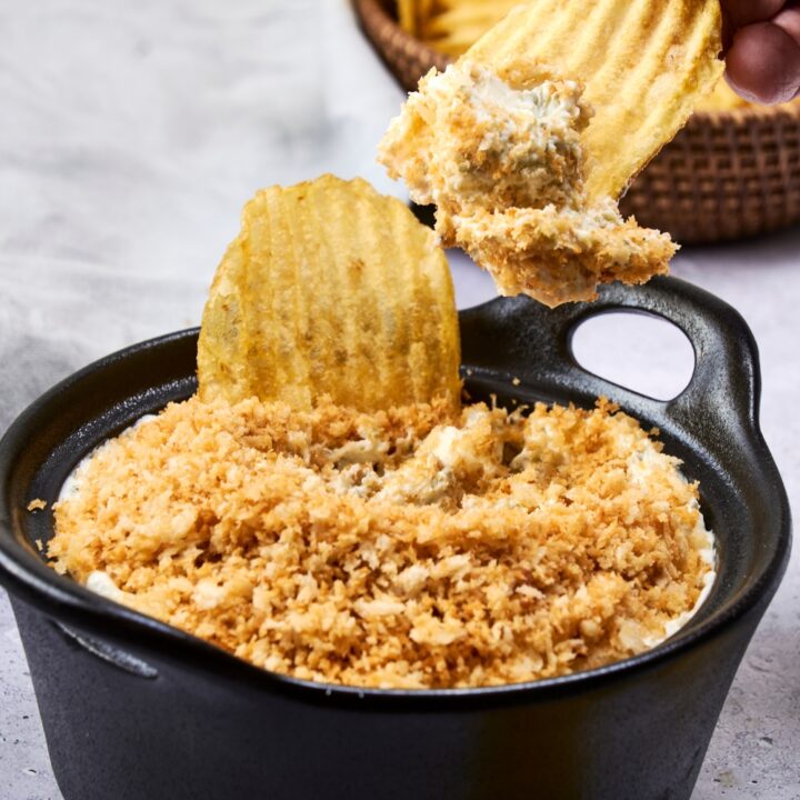 A serving bowl with fried pickle dip topped with toasted panko crumbs. Potato chips are being dipped into it.