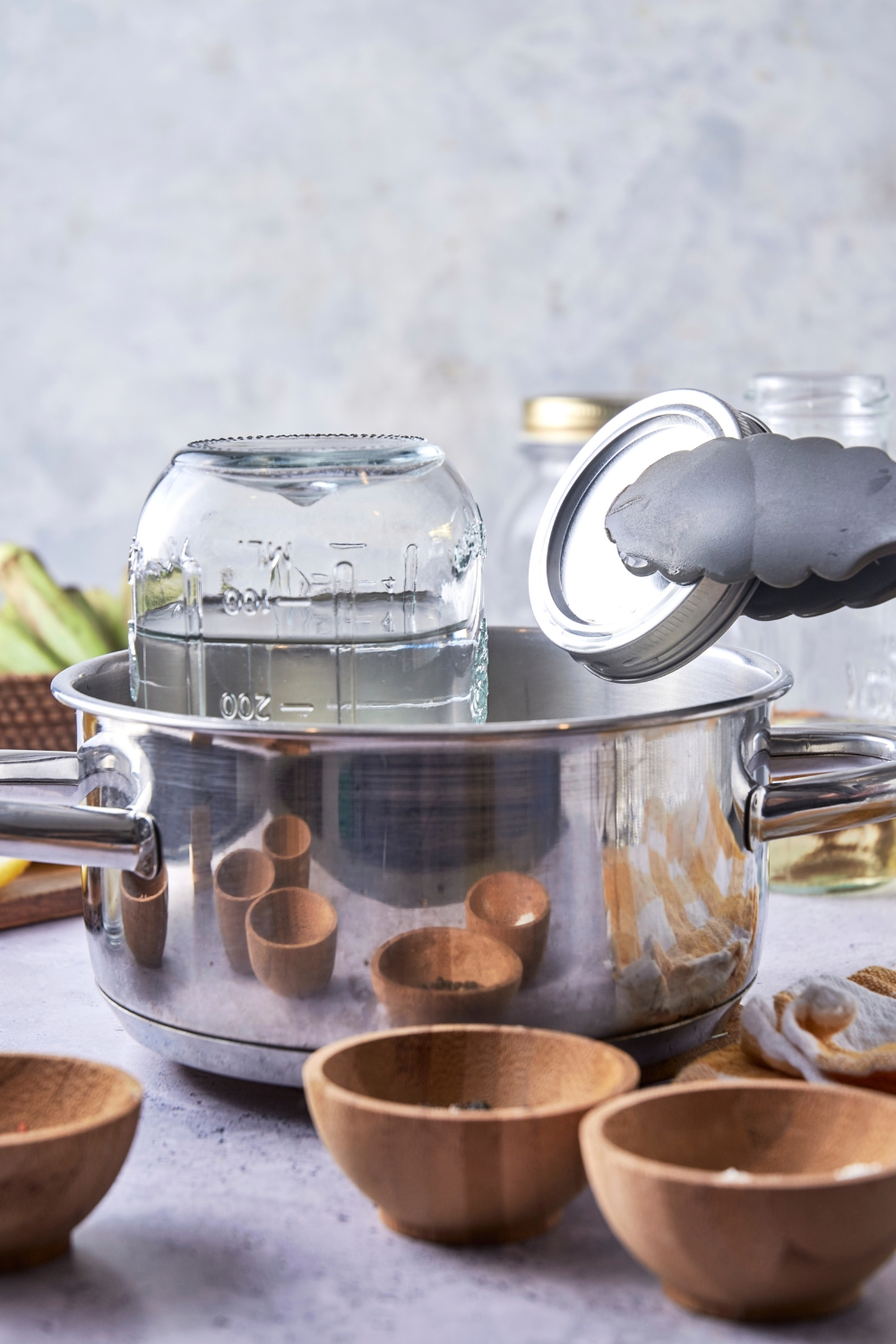 A pot with the jar upside down in it. Tongs are holding the lid.