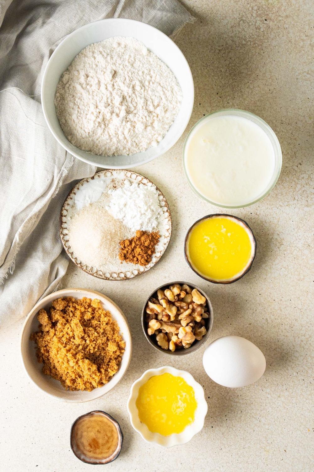 A countertop with multiple bowls with various ingredients like flour, sugar, cinnamon, baking powder, baking soda, melted butter, chopped walnuts, brown sugar, rum, buttermilk, and an egg.