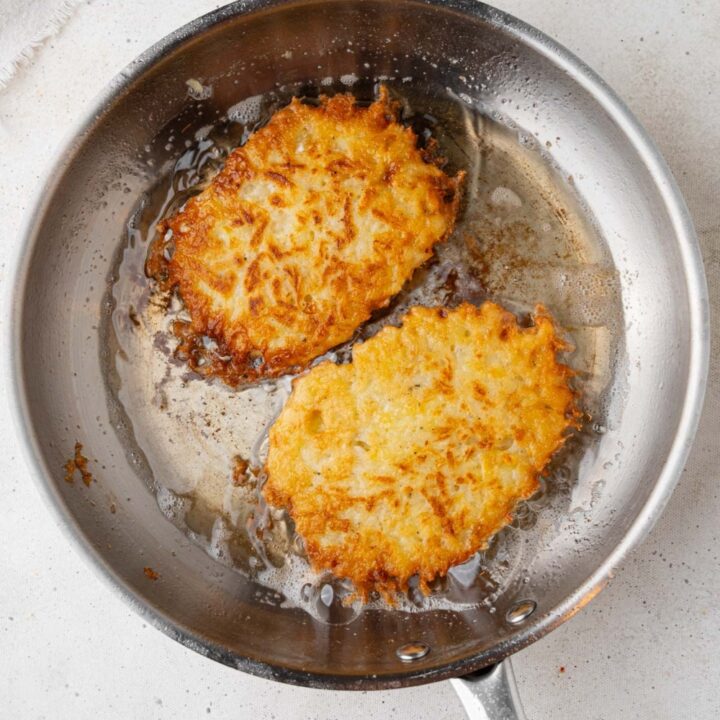 A skillet with two copycat McDonald's hashbrown patties frying in oil. The skillet is on a marble counter with a white cloth napkin off to the side.