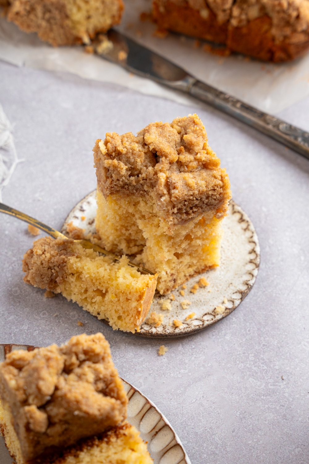 A square slice of Bisquick coffee cake on a ceramic plate with a fork slicing a bite. The plate is on a white counter with an additional plate of coffee cake.