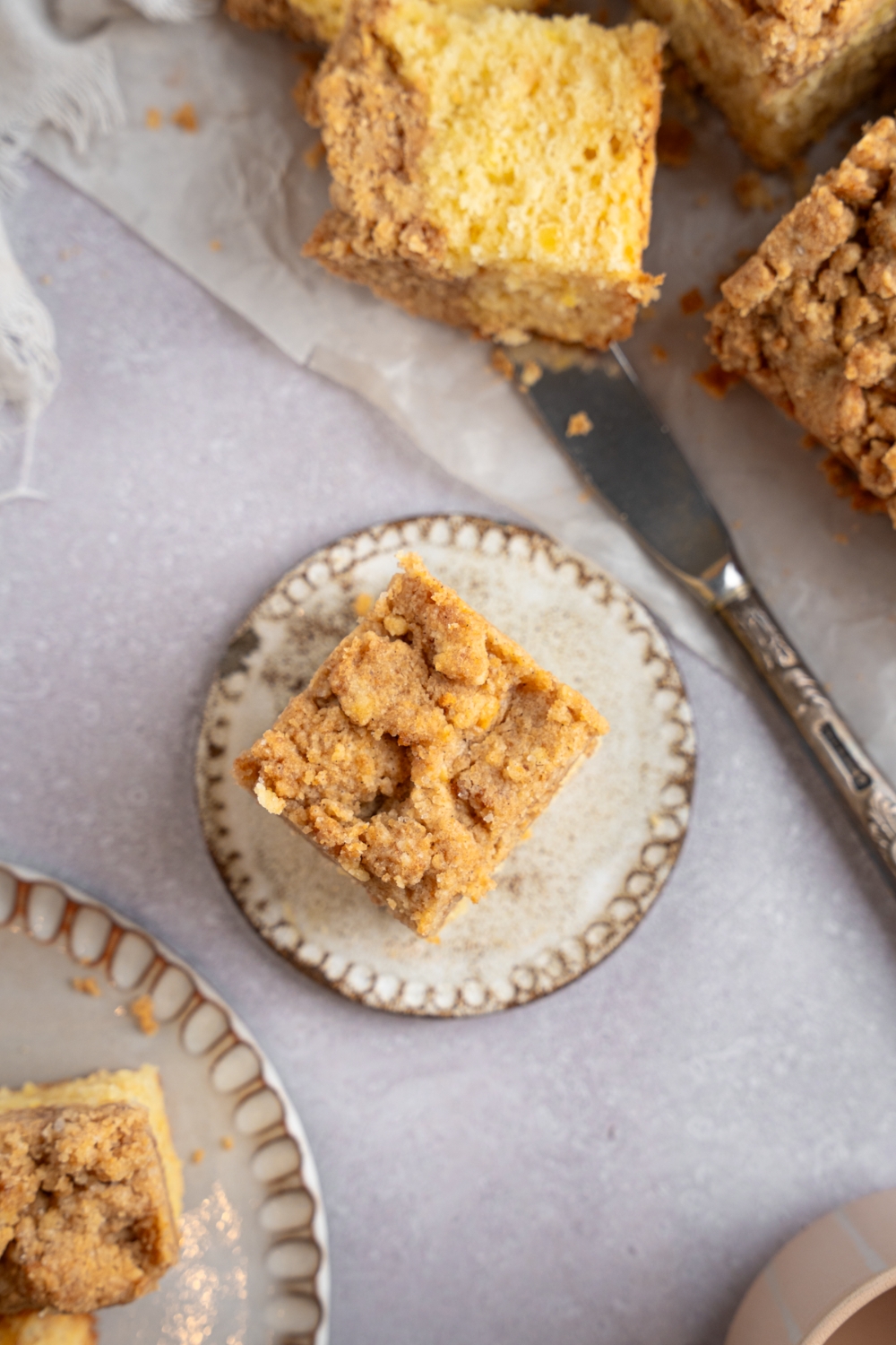 A square slice of Bisquick coffee cake on a ceramic plate. The plate is on a white counter with additional slices of cake on a sheet of parchment paper. There is a knife next to the coffee cake.
