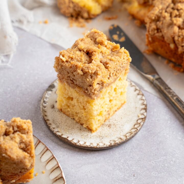 A square slice of Bisquick coffee cake on a ceramic plate. The plate is on a white counter with additional slices of cake on a sheet of parchment paper. There is a knife next to the coffee cake.