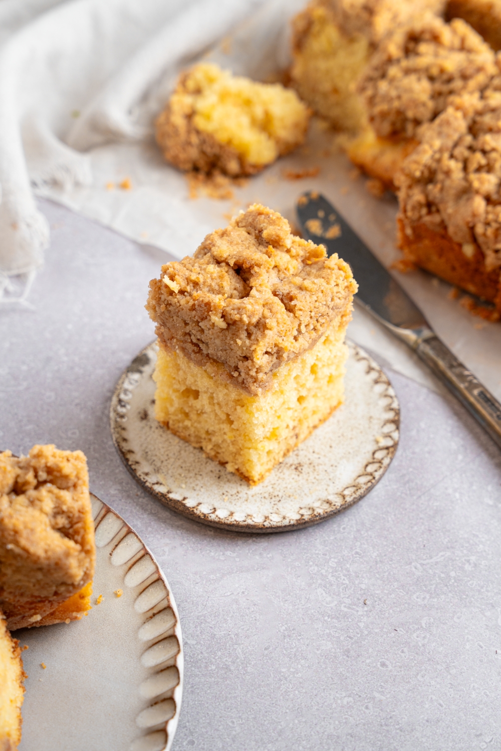 A square slice of Bisquick coffee cake on a ceramic plate. The plate is on a white counter with additional slices of cake on a sheet of parchment paper. There is a knife next to the coffee cake.