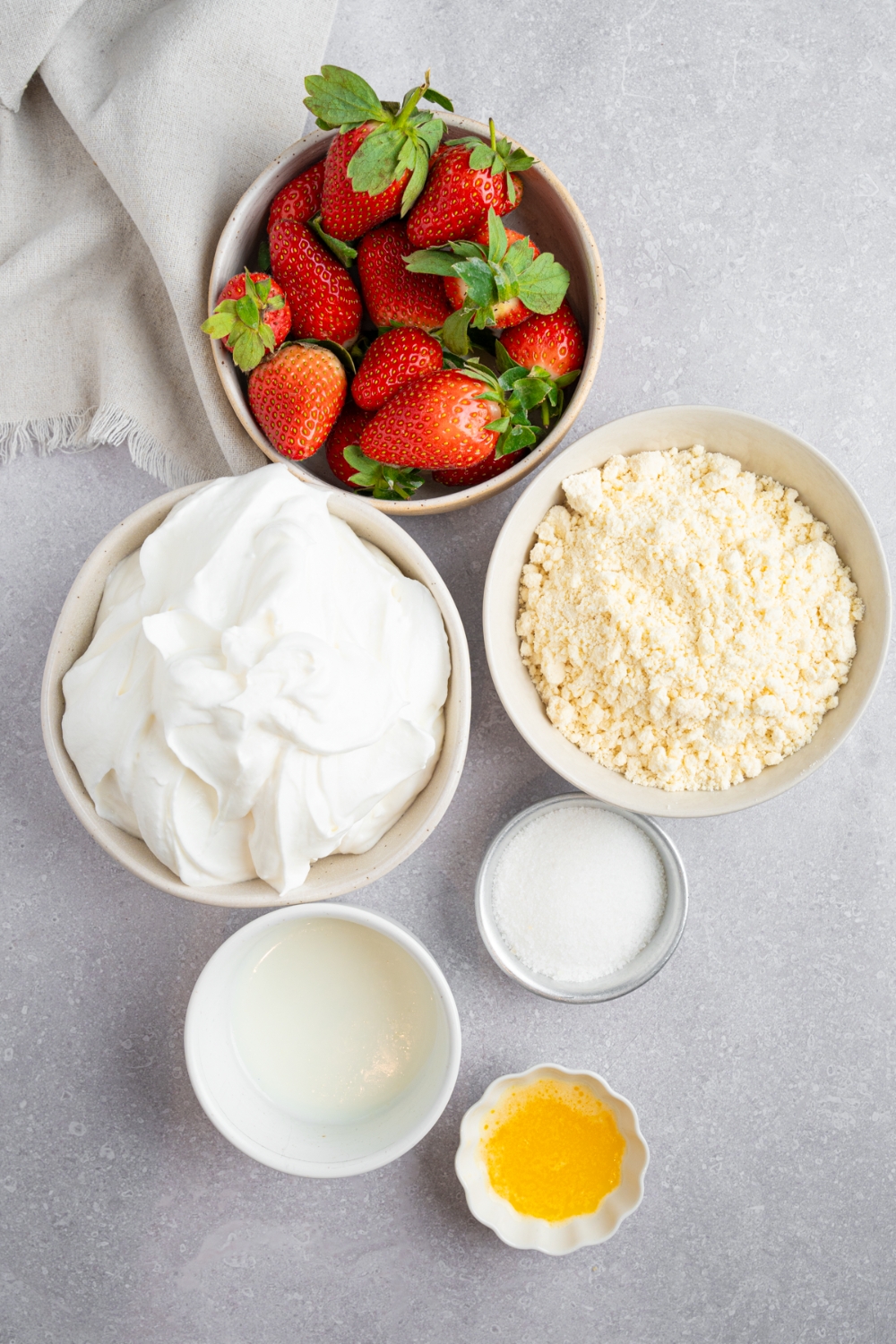 An overhead shot of several bowls in various sizes containing ingredients to make Bisquick strawberry shortcake including Bisquick baking mix, whipped cream, fresh strawberries, milk, sugar, and melted butter.