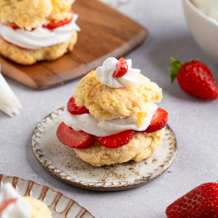 A Bisquick strawberry shortcake served on a small ceramic plate next to a wooden server with additional strawberry shortcakes. The plate is on a white counter with fresh strawberries.