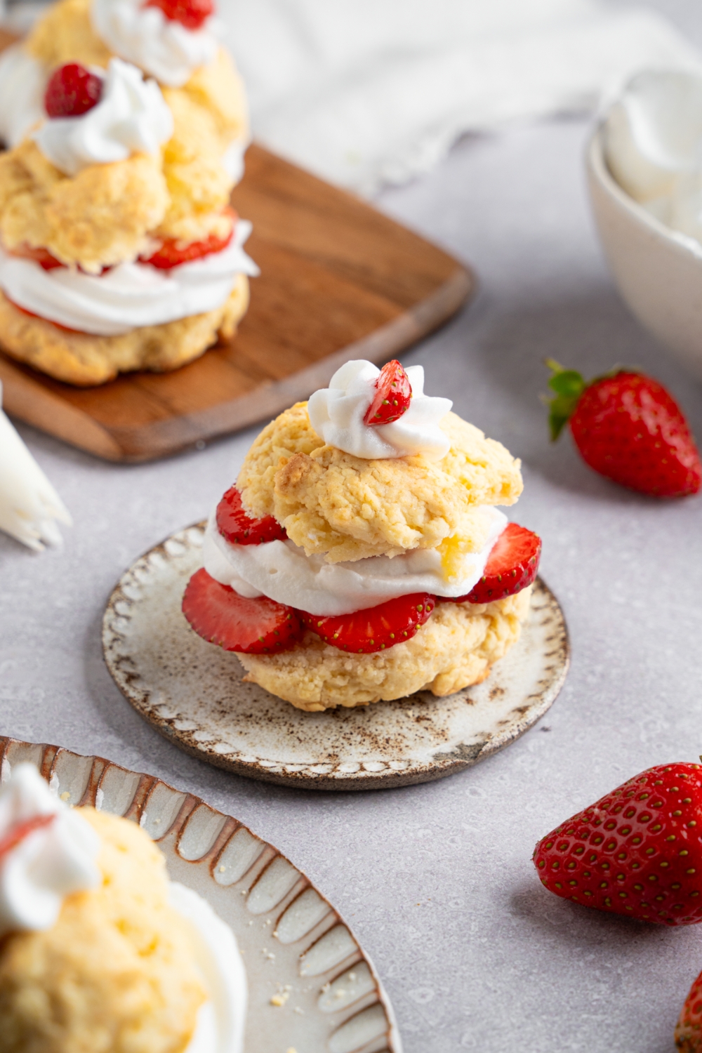 A Bisquick strawberry shortcake served on a small ceramic plate next to a wooden server with additional strawberry shortcakes. The plate is on a white counter with fresh strawberries.