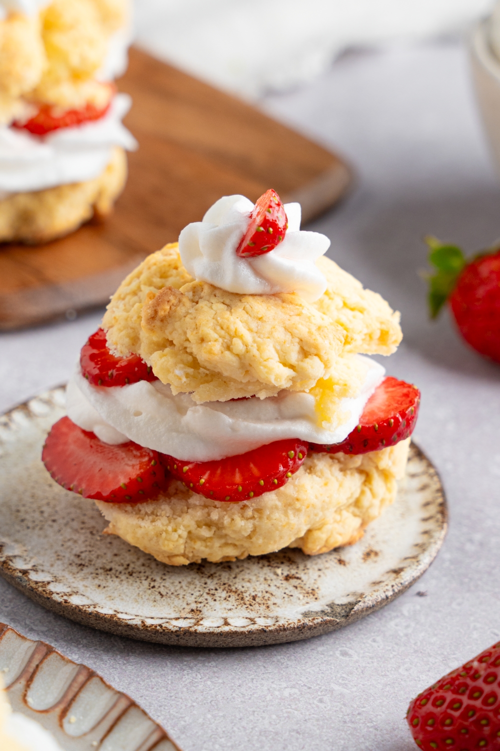 A Bisquick strawberry shortcake served on a small ceramic plate next to a wooden server with additional strawberry shortcakes. The plate is on a white counter with fresh strawberries.