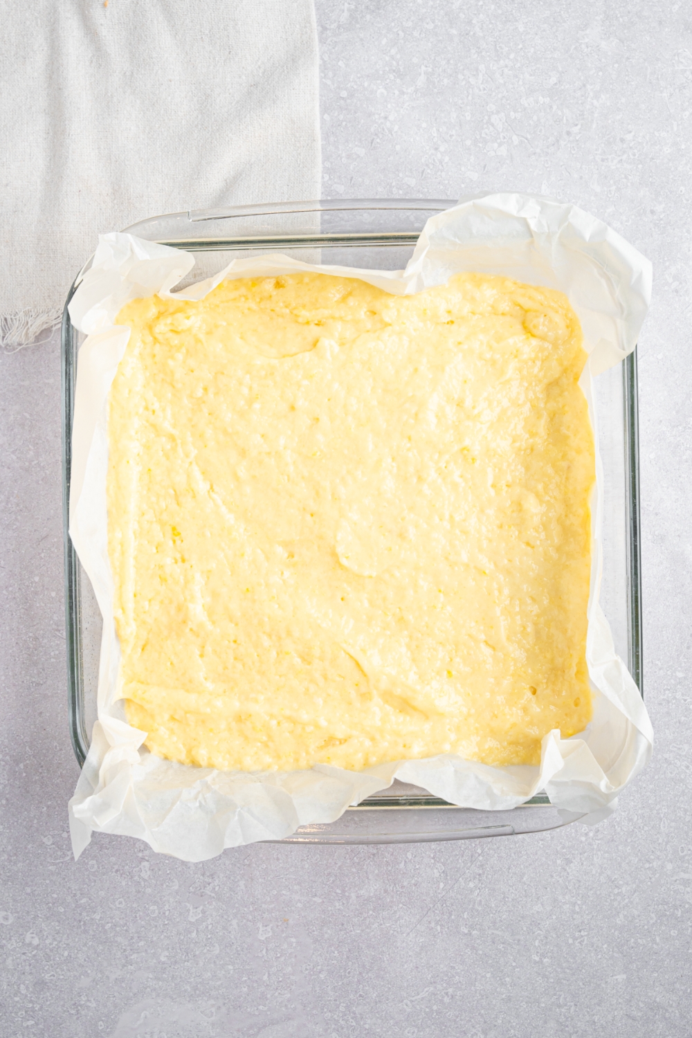 A square glass baking dish lined with parchment paper and filled with Bisquick dough. The dish is on a white counter with a white cloth napkin.
