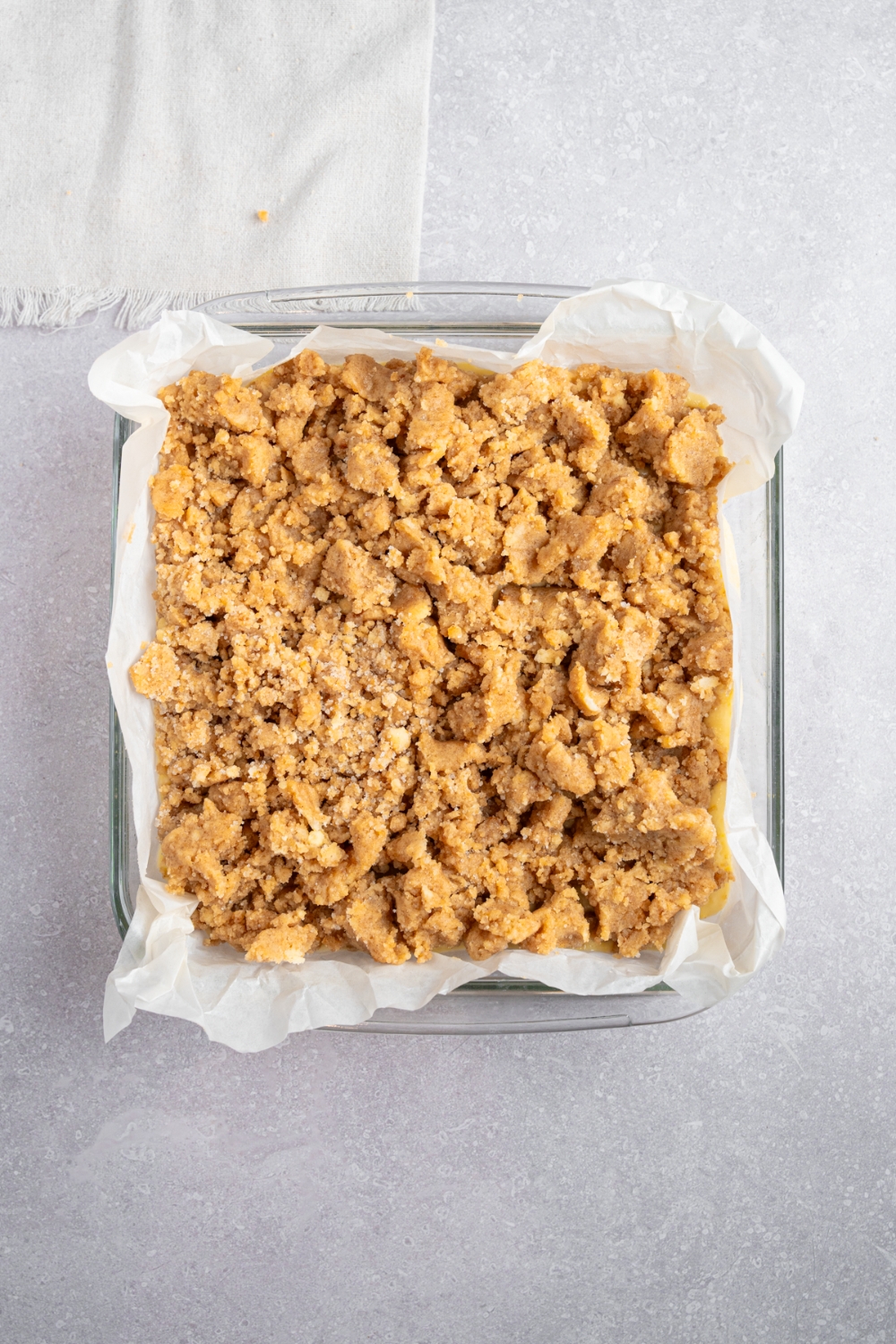 Uncooked Bisquick coffee cake in a square glass baking dish lined with parchment paper. The dish is on a white counter with a white cloth napkin.