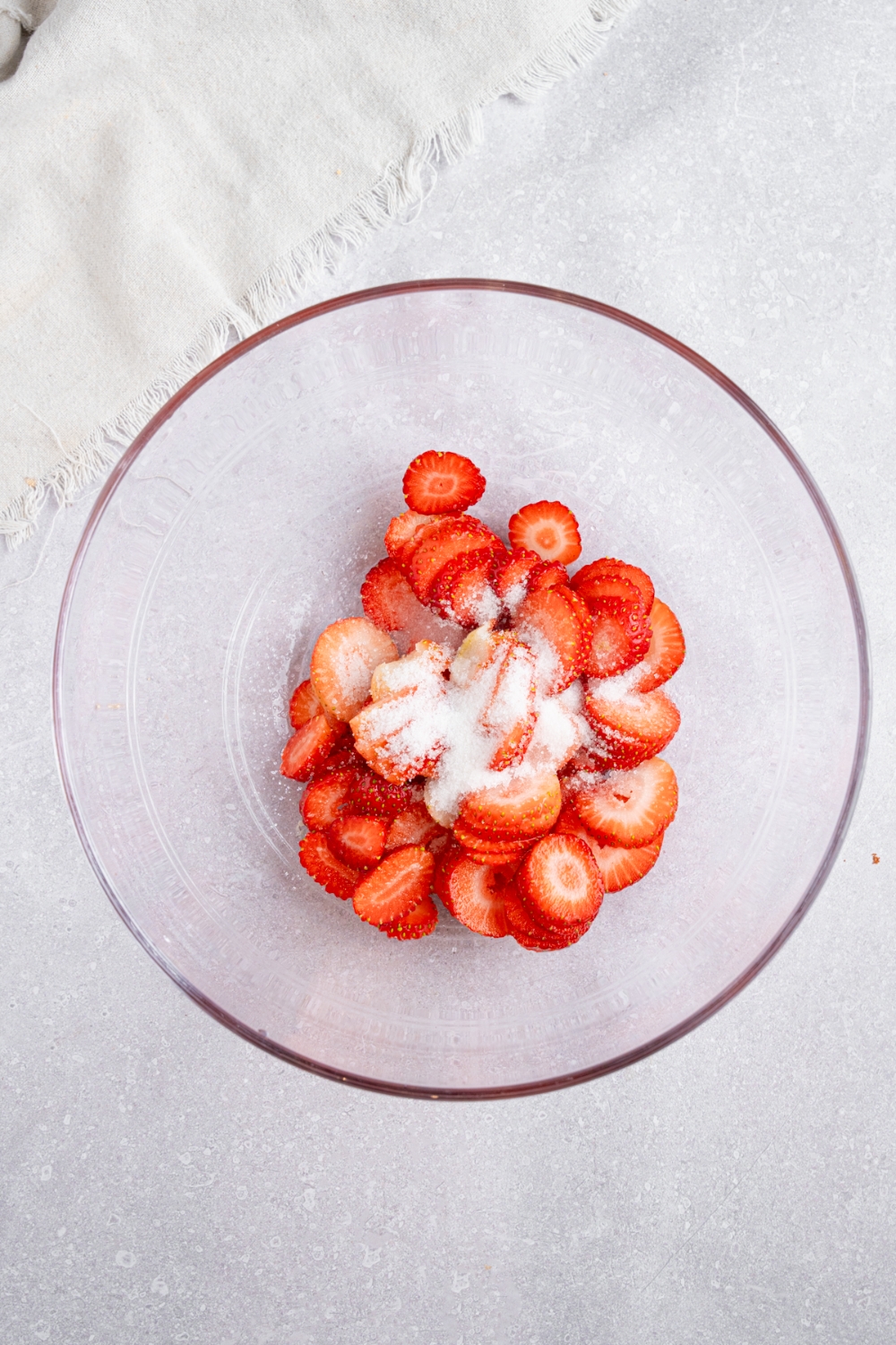 A glass bowl with sliced strawberries topped with sugar. The bowl is on a white counter with a white cloth napkin.