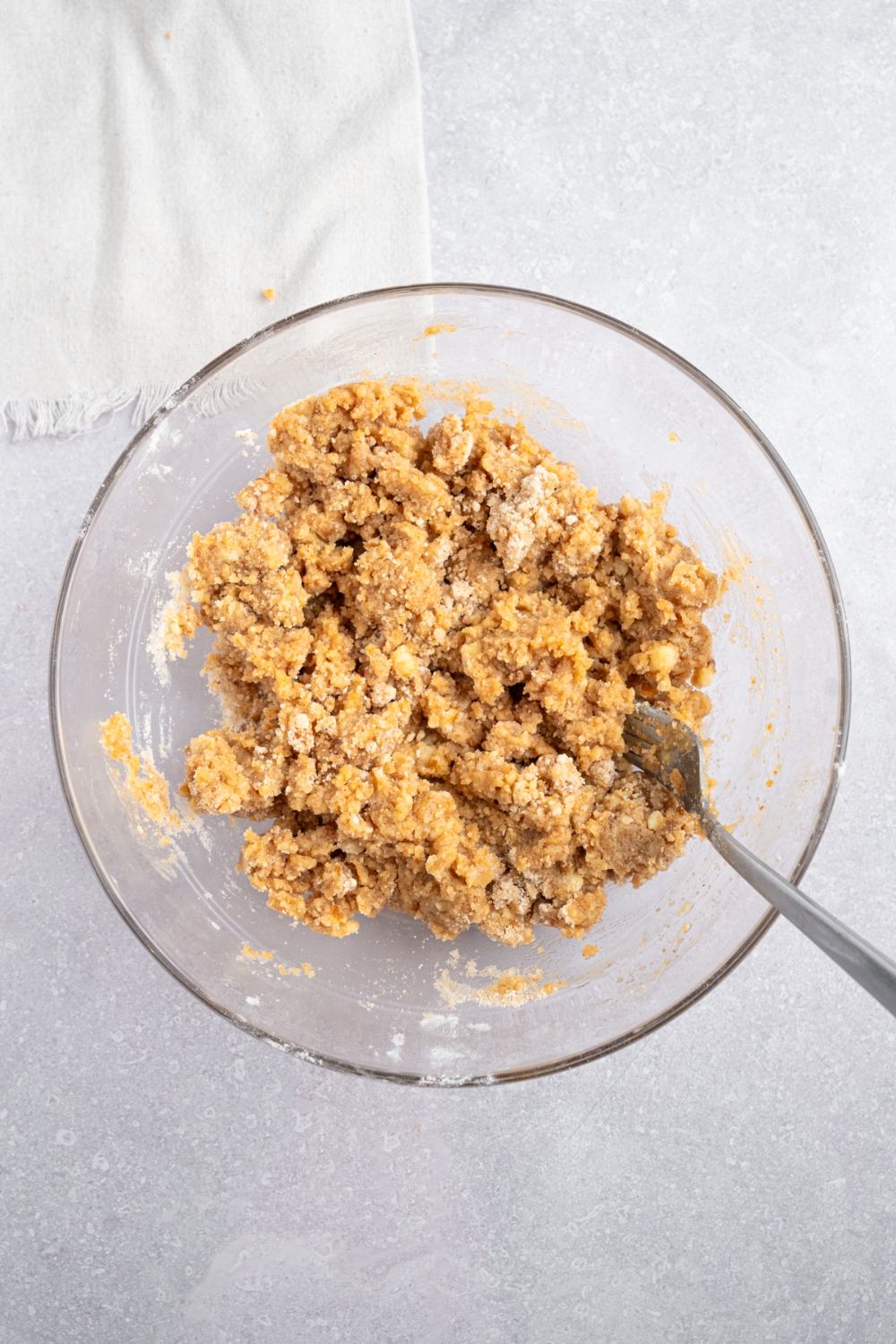 A glass bowl with a fork stirring a crumb topping mixture including brown sugar, butter, Bisquick mix, and cinnamon. The bowl is on a white counter with a white napkin.