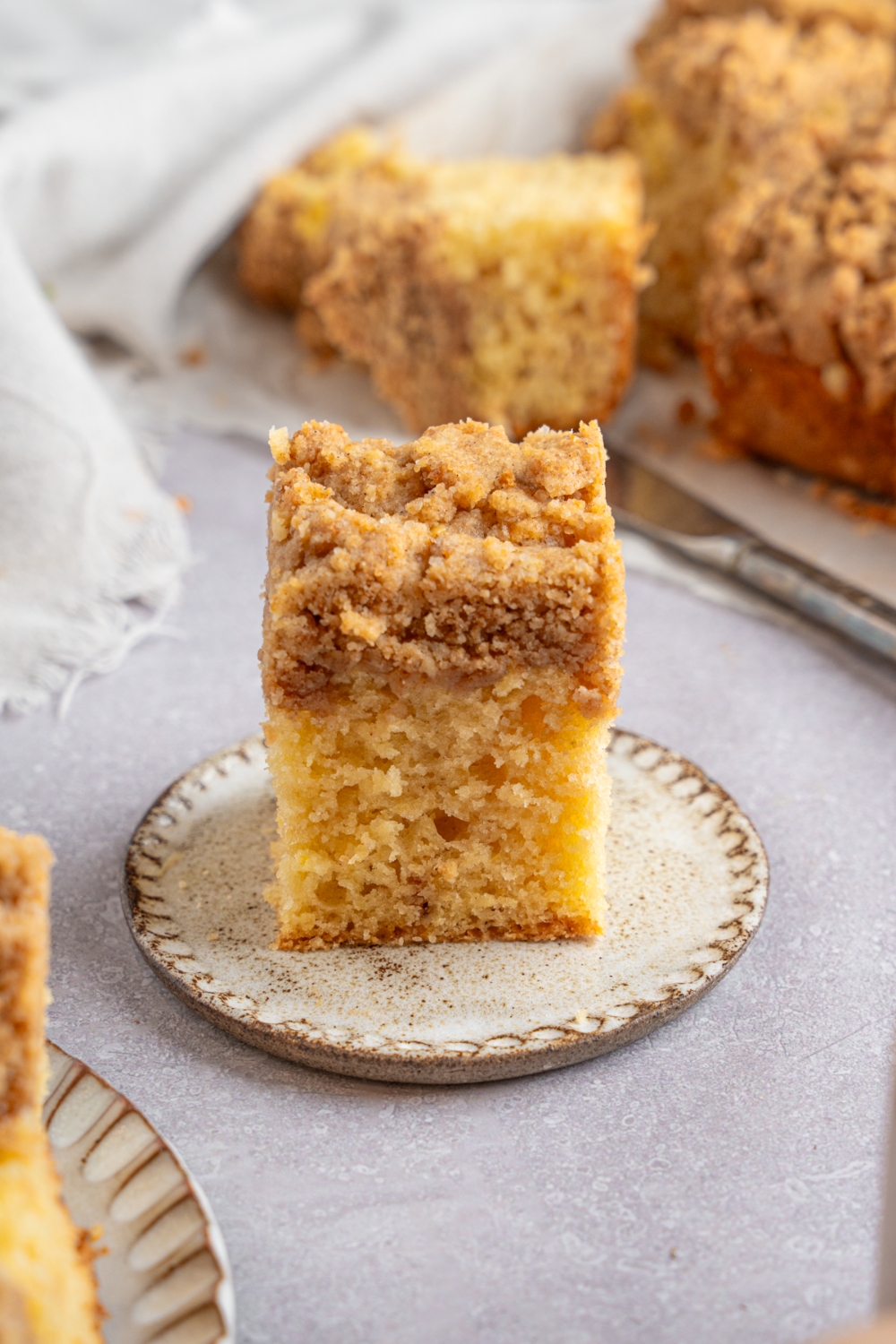 A square slice of Bisquick coffee cake on a ceramic plate. The plate is on a white counter with additional slices of cake on a sheet of parchment paper. There is a knife next to the coffee cake.