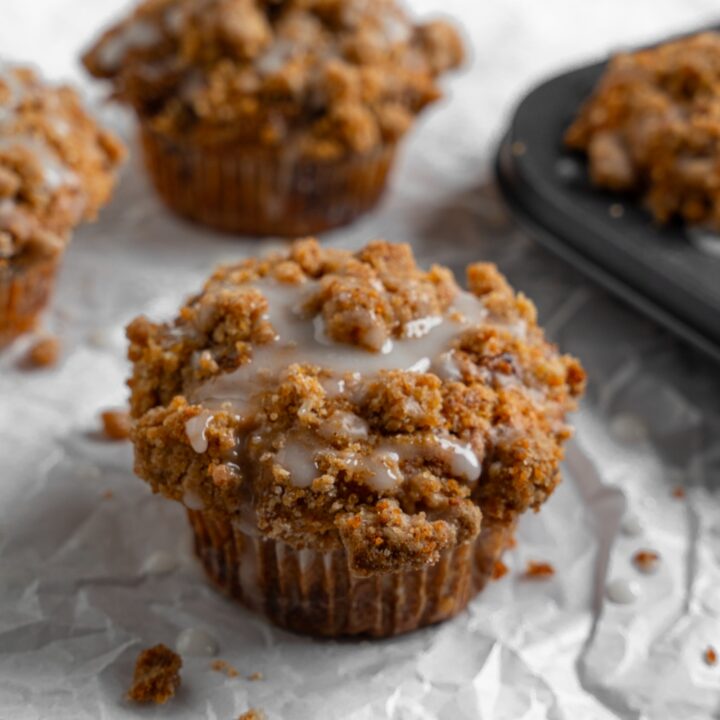 Coffee cake muffins on a sheet of parchment paper with a tray of baked muffins. The muffins are drizzled with a vanilla glaze.