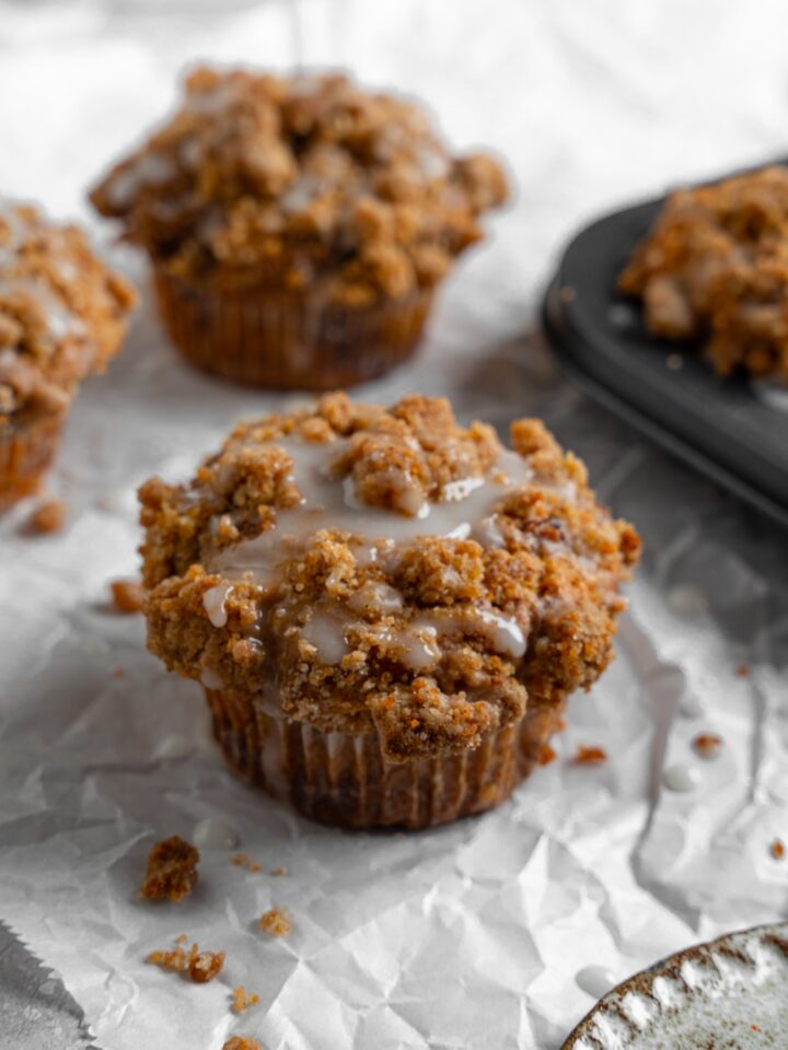 Coffee cake muffins on a sheet of parchment paper with a tray of baked muffins. The muffins are drizzled with a vanilla glaze.