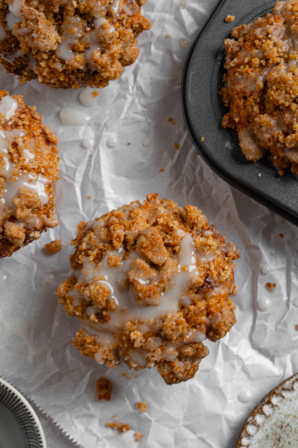 Coffee cake muffins on a sheet of parchment paper with a tray of baked muffins. The muffins are drizzled with a vanilla glaze.