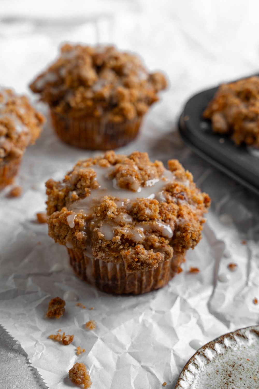 Coffee cake muffins on a sheet of parchment paper with a tray of baked muffins. The muffins are drizzled with a vanilla glaze.
