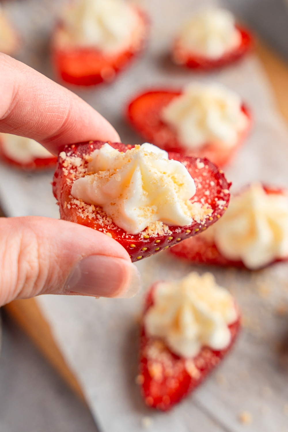 A close up of a hand holding a sliced deviled strawberry with cheesecake filling on top. In the background is a board lined with parchment paper with additional slices of deviled strawberries garnished with graham cracker crumbs.