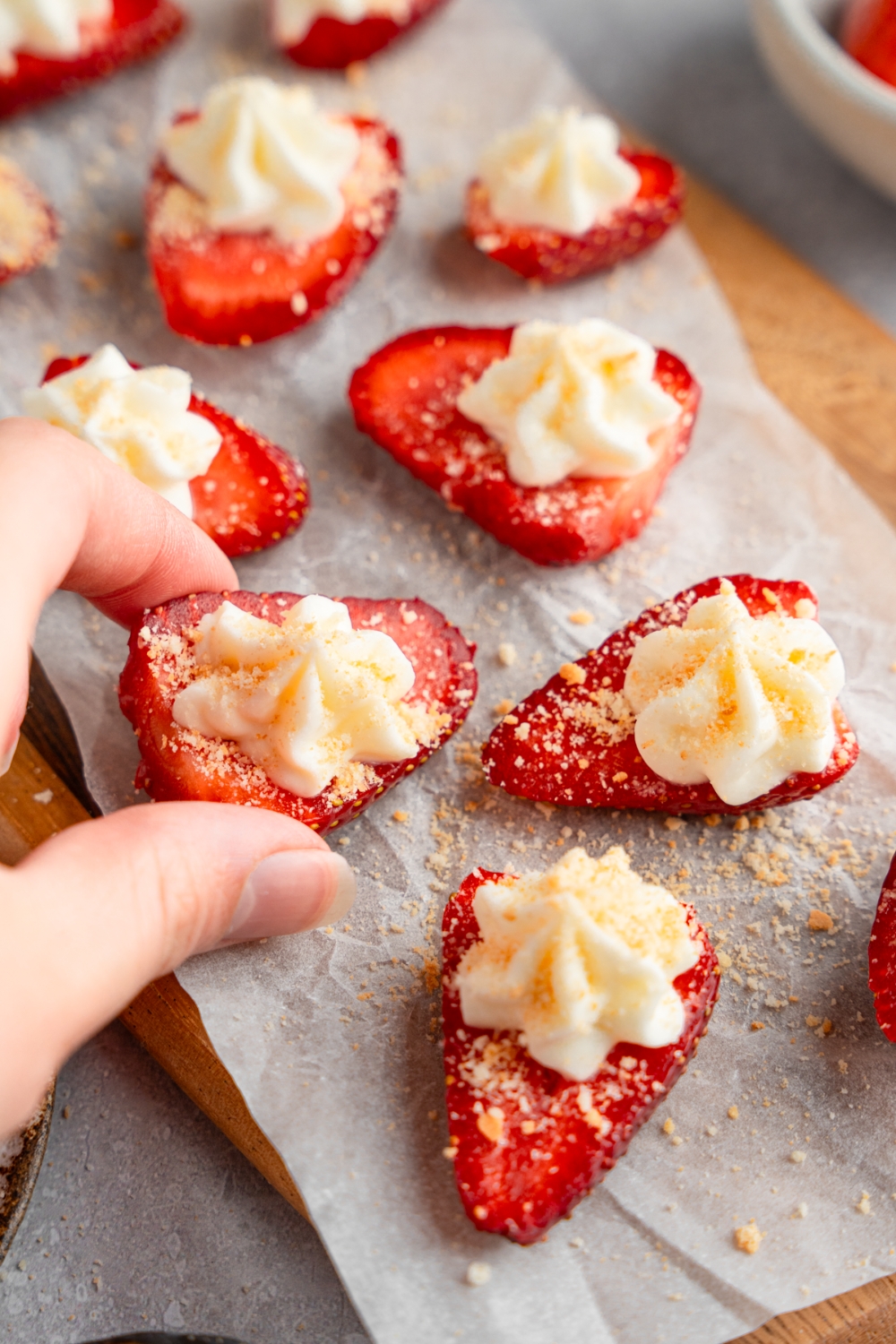 A wooden board lined with parchment paper with several sliced of deviled strawberries topped with cheesecake filling and garnished with graham cracker crumbs. A hand is taking a strawberry.