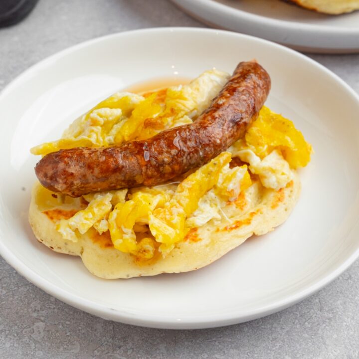 A pancake taco on a white plate. The pancake is layered with scrambled egg and a sausage link drizzled with syrup. The plate is on a white counter with an additional pancake.