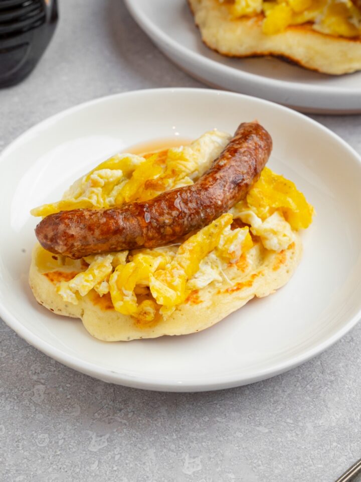 A pancake taco on a white plate. The pancake is layered with scrambled egg and a sausage link drizzled with syrup. The plate is on a white counter with an additional pancake.