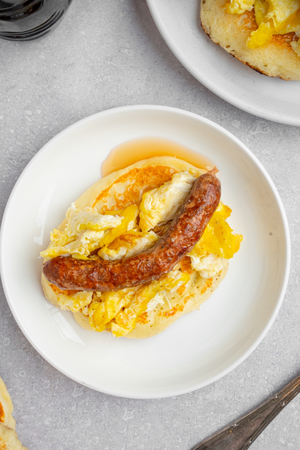 A pancake taco on a white plate. The pancake is layered with scrambled egg and a sausage link drizzled with syrup. The plate is on a white counter with an additional pancake.