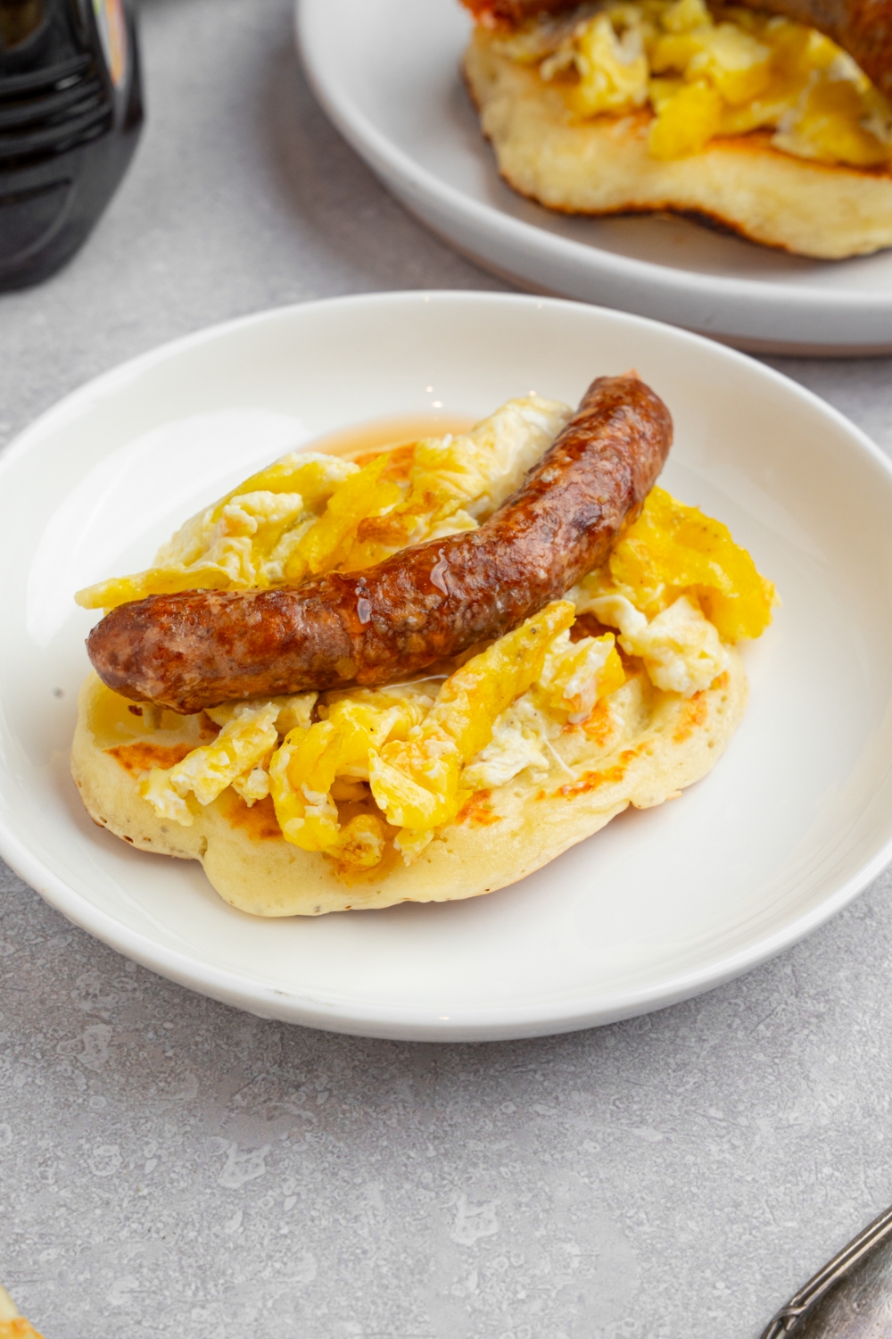A pancake taco on a white plate. The pancake is layered with scrambled egg and a sausage link drizzled with syrup. The plate is on a white counter with an additional pancake.