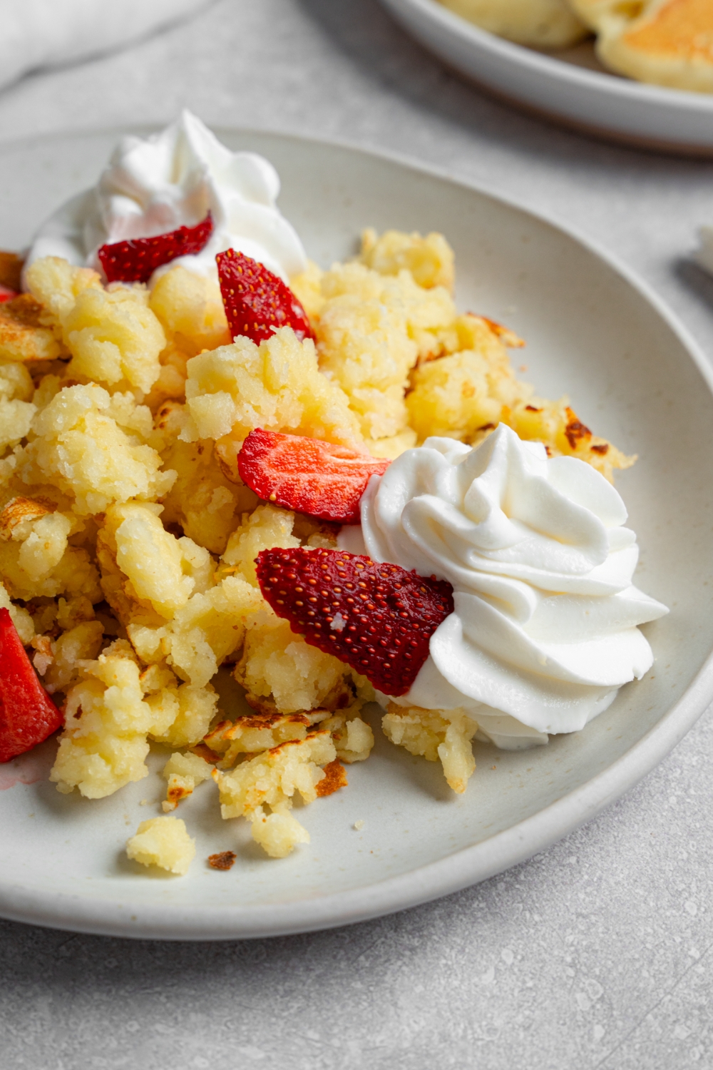 A white plate with scrambled pancakes with strawberries and whipped cream. The plate is on a white counter with a plate of pancakes.