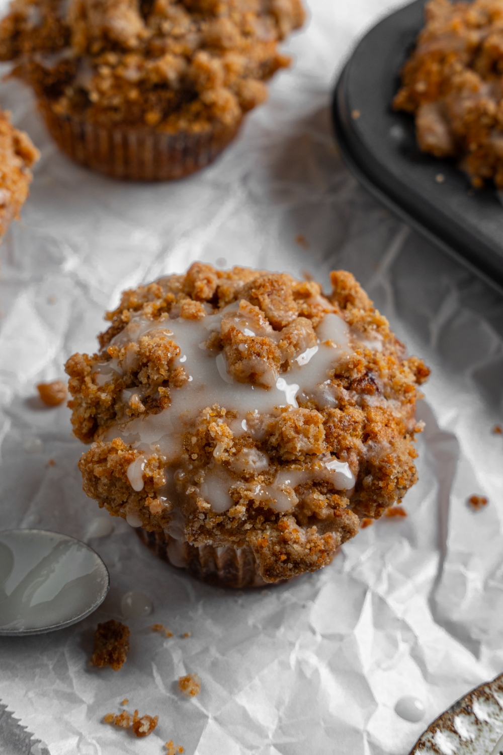 Coffee cake muffins on a sheet of parchment paper with a tray of baked muffins. The muffins are drizzled with a vanilla glaze.