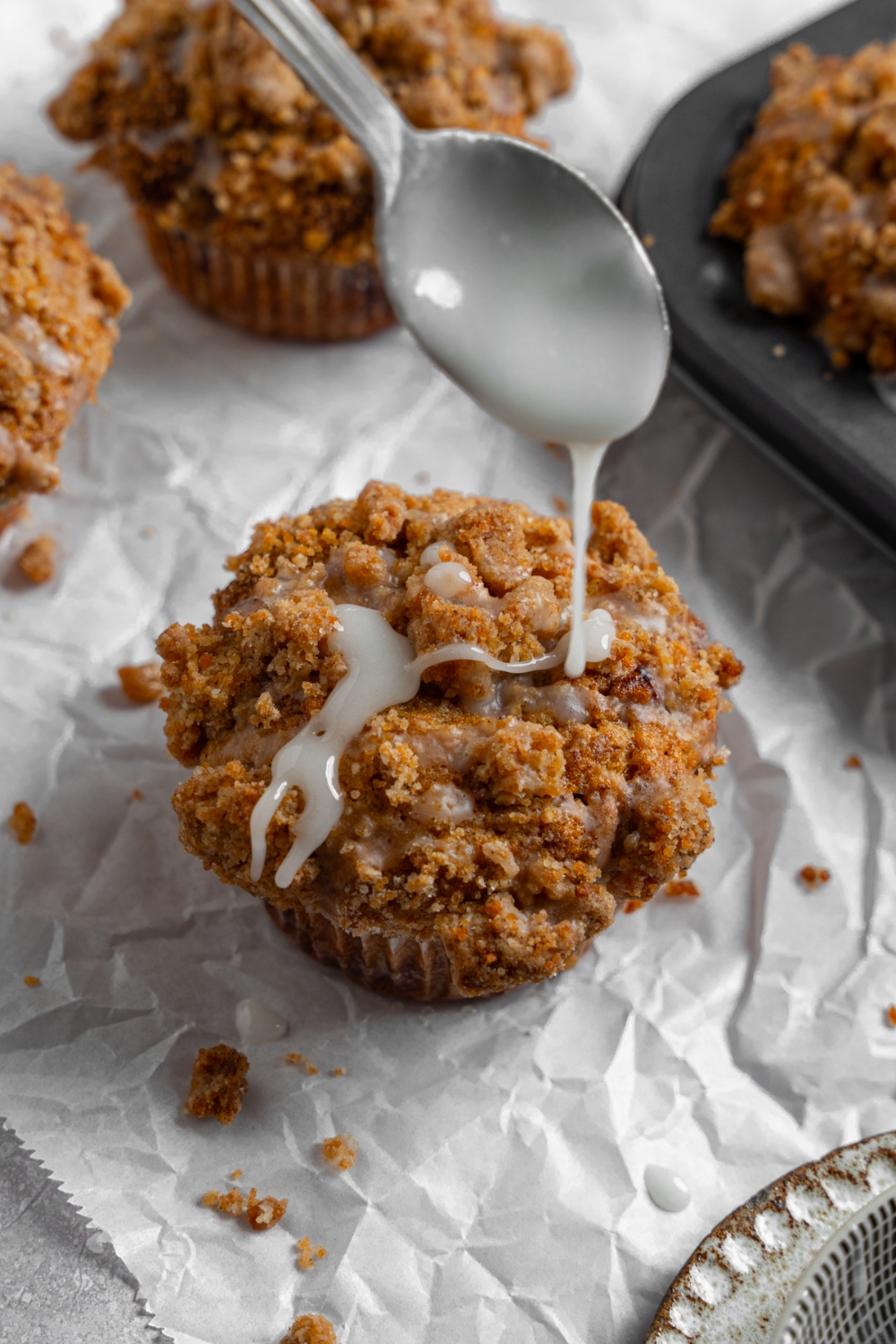 Coffee cake muffins on a sheet of parchment paper with a tray of baked muffins. A spoon is drizzling a vanilla glaze over the muffin.