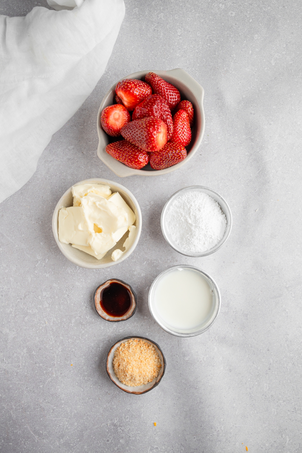 An overhead shot of several bowls in various sizes containing ingredients to make deviled strawberries including strawberries, cream cheese, vanilla, powdered sugar, whipping cream, and graham cracker crumbs.