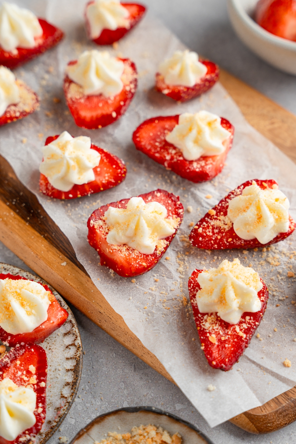 A wooden board lined with parchment paper with several sliced of deviled strawberries topped with cheesecake filling and garnished with graham cracker crumbs. The board is on a white counter with a bowl of strawberries.