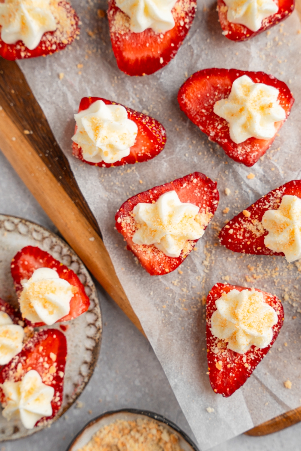 A wooden board lined with parchment paper with several sliced of deviled strawberries topped with cheesecake filling and garnished with graham cracker crumbs. The board is on a white counter with an additional plate of strawberries.