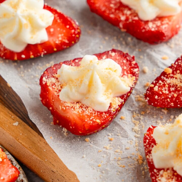 A wooden board lined with parchment paper with several sliced of deviled strawberries topped with cheesecake filling and garnished with graham cracker crumbs.
