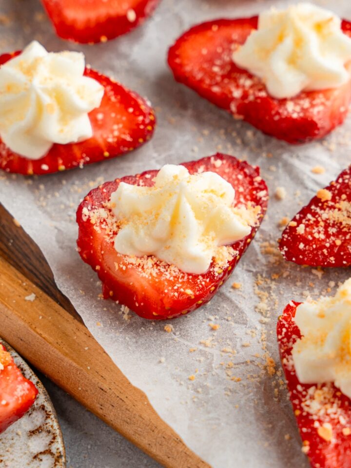 A wooden board lined with parchment paper with several sliced of deviled strawberries topped with cheesecake filling and garnished with graham cracker crumbs.