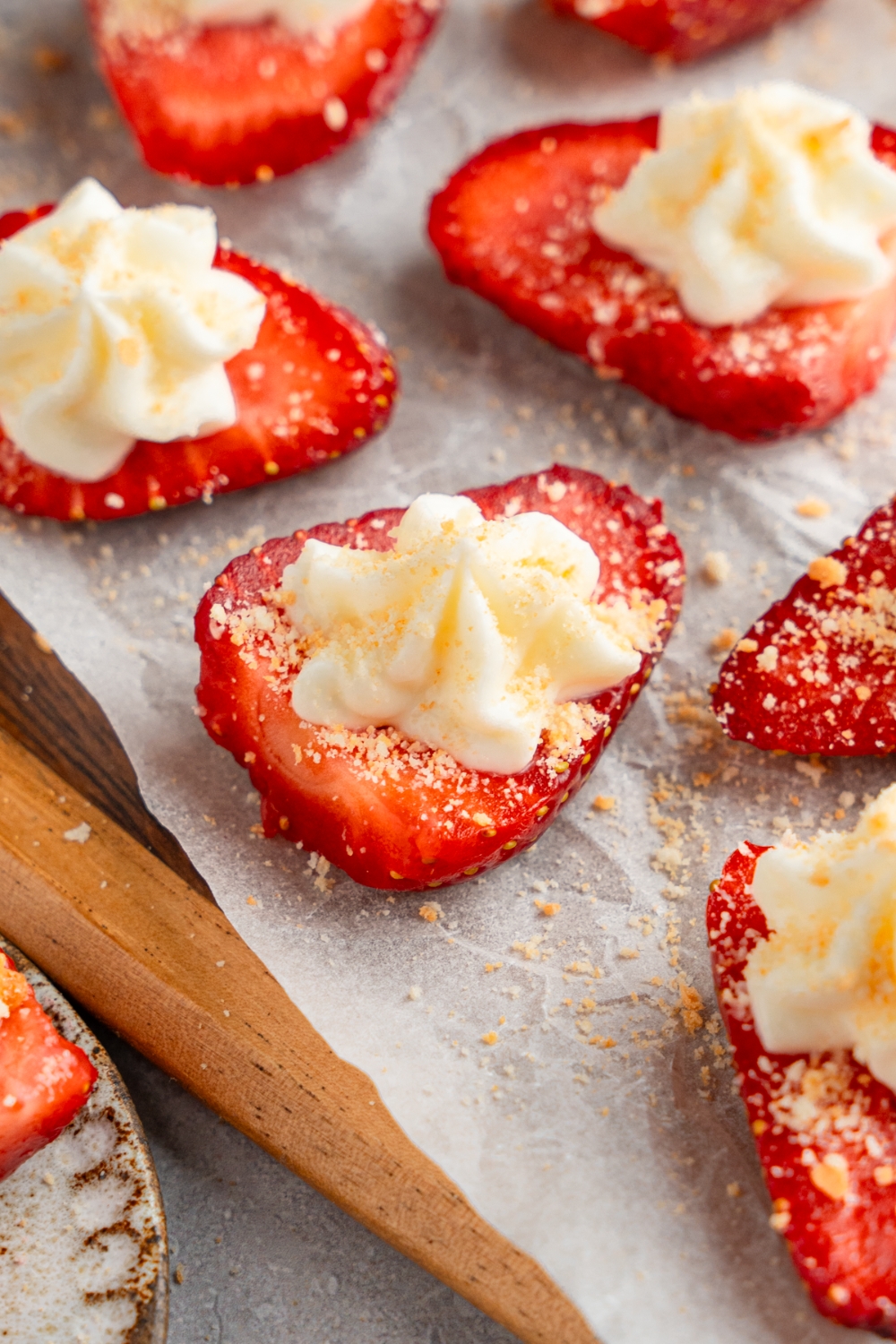 A wooden board lined with parchment paper with several sliced of deviled strawberries topped with cheesecake filling and garnished with graham cracker crumbs.
