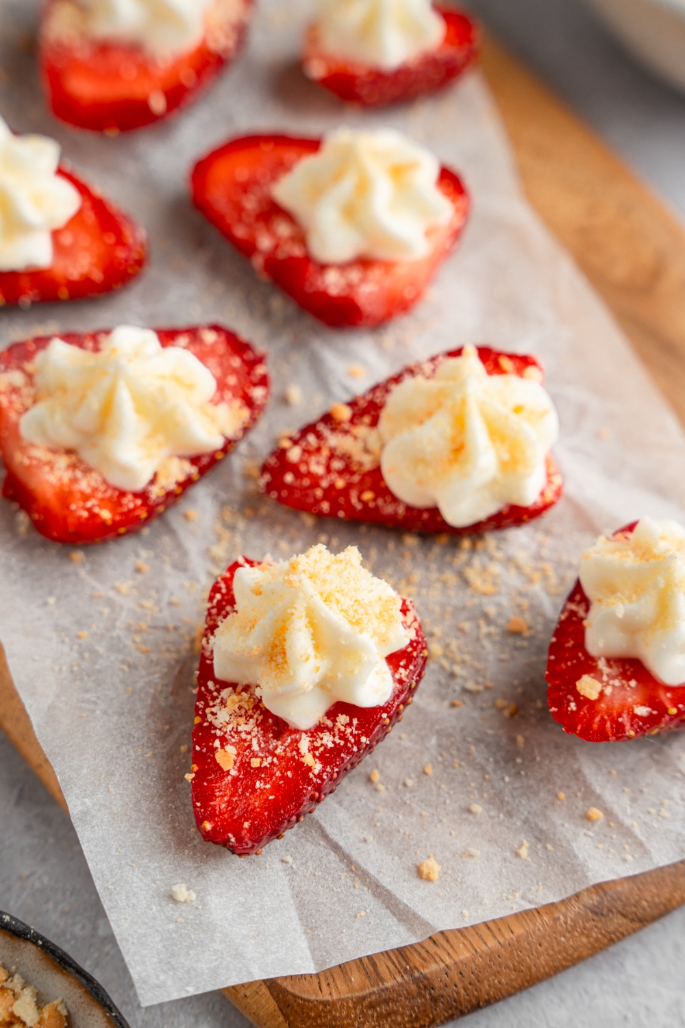 A wooden board lined with parchment paper with several sliced of deviled strawberries topped with cheesecake filling and garnished with graham cracker crumbs.