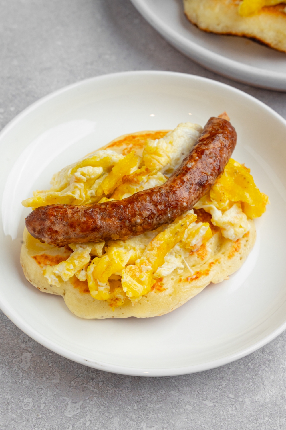 A pancake taco on a white plate. The pancake is layered with scrambled egg and a sausage link drizzled with syrup. The plate is on a white counter with an additional pancake.