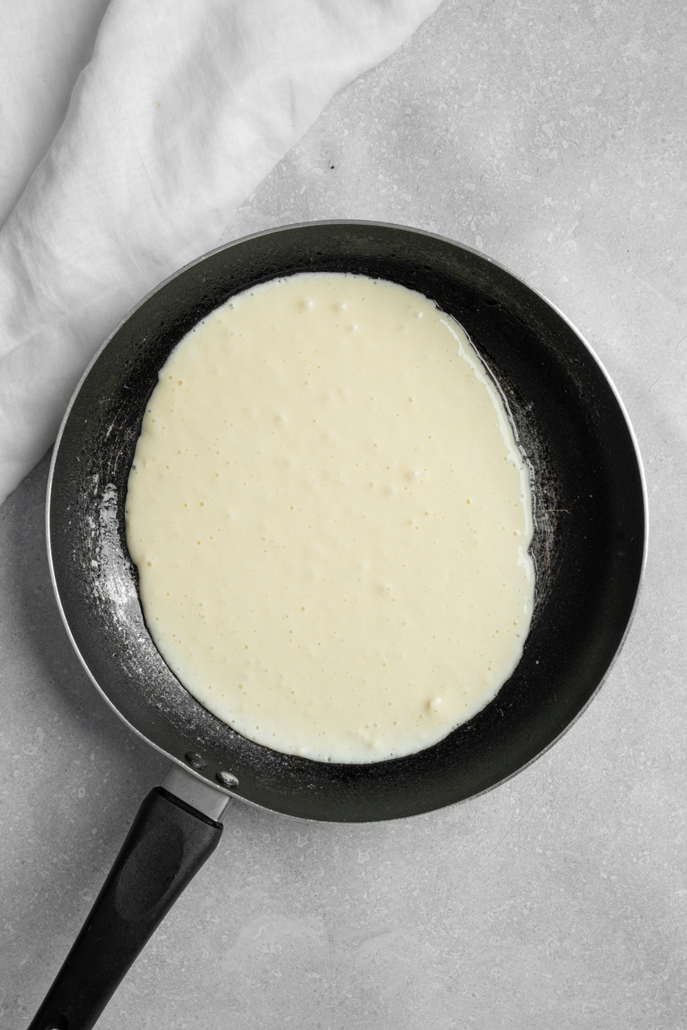 A skillet with an uncooked pancake. The skillet is on a white counter with a white cloth napkin.