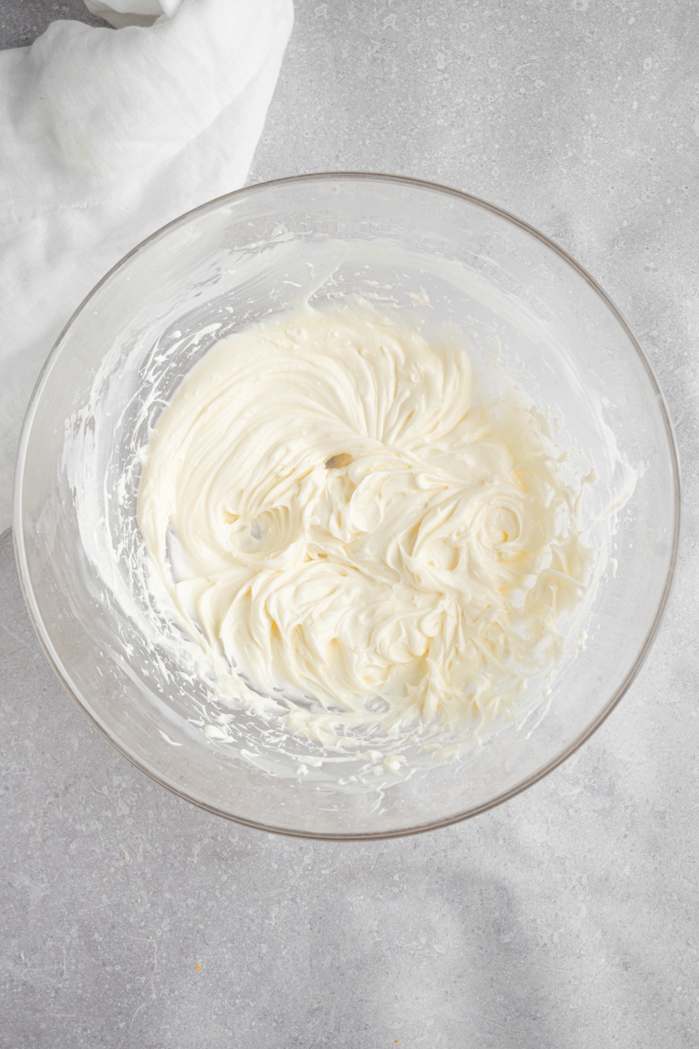 A glass bowl with whipped cream cheese, sugar, and vanilla. The bowl is on a white counter with a white cloth napkin.