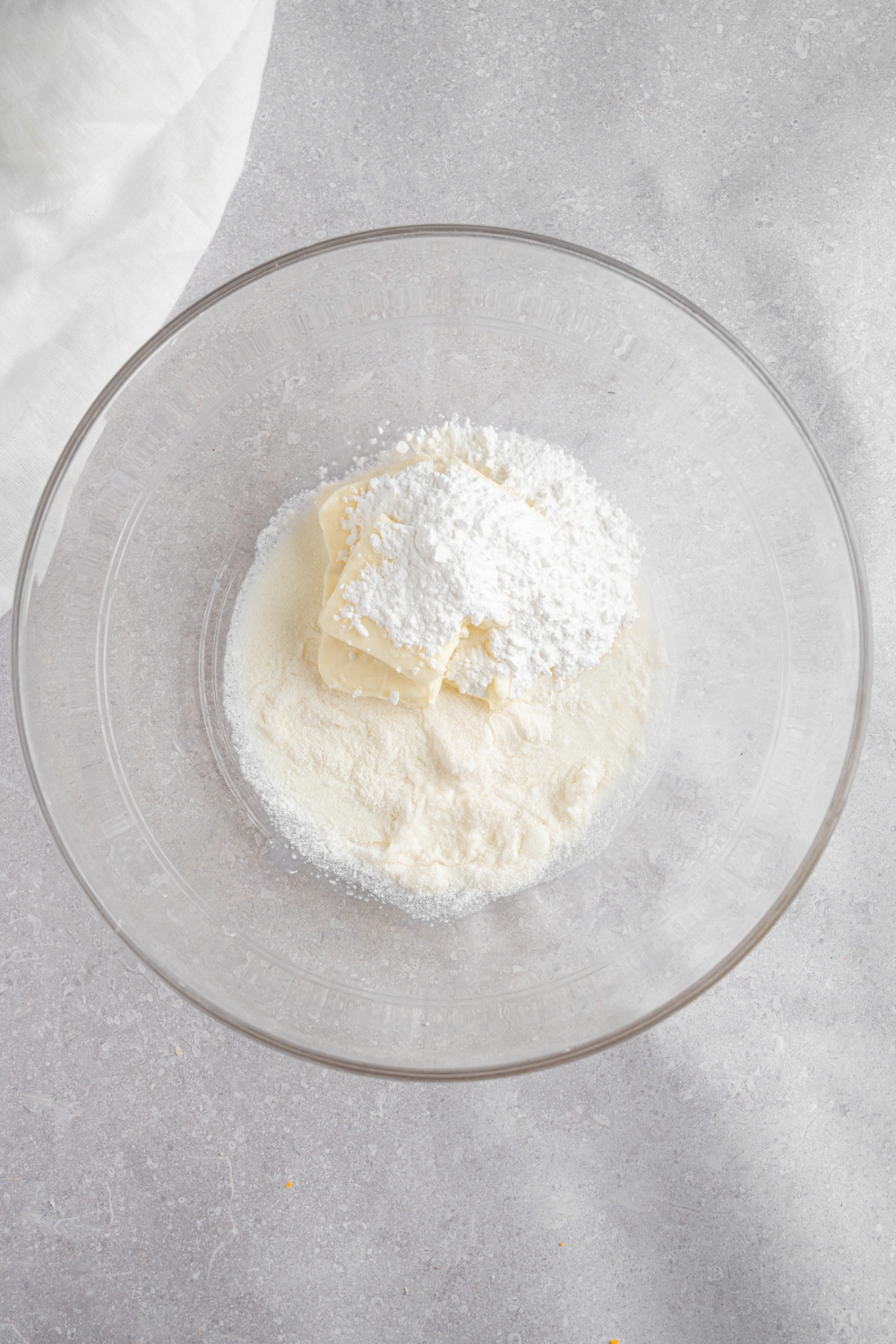 A glass bowl with cream cheese, heavy whipping cream, and powdered sugar. The bowl is on a white counter with a white cloth napkin.